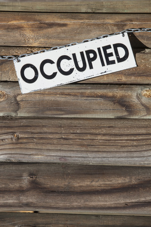 white metal sign with the word occupied hanging at a chain  in front of a wooden wall. Concept for all things being occupiedの写真素材