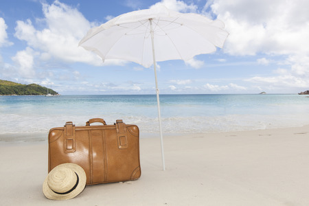 travel concept with a vintage brown suitcase and a straw sunhat under a white sunshade at a tropical beach with a turquoise sea, Seychelles, Africaの写真素材
