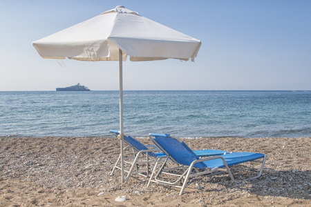 white sunshade and blue sunbeds at pebble beach with a luxury yacht at the horizon, Latsi beach, Cyprus, Greece, Europeの写真素材