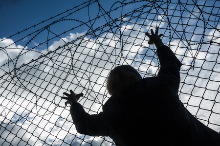 black silhouette of a refugee at a border fence with barbwire, grabbing the mesh wire, social concept for asylum, illegal immigration, escape and prisonの写真素材