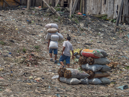 Iquitos, Peru - Sep 25, 2018: Men are carring the bags with charcoal. A huge pollution can be seen in the background on the bank of Itaya River. Low water season of Amazon. BelÃ©n district of Iquitos, Peruのeditorial素材