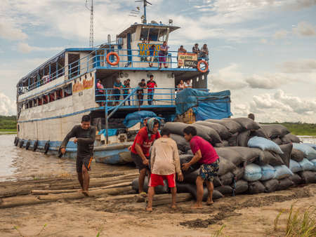 Amazon River, Peru - December 04, 2018: Cargo boat and the workers collecting the bags with rice. Rice fields on the island on the Amazon River during the low water season. South Americaのeditorial素材