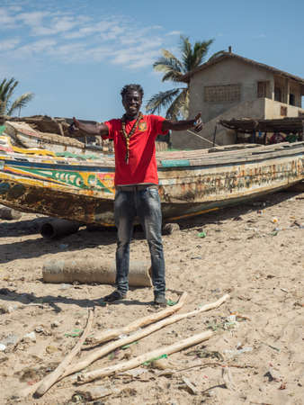 Nianing, Senegal - January 24, 2019: Smiling Senegalese man on the background of colorful wooden fishing boats standing on the sandy beach in Senegal. Africaのeditorial素材