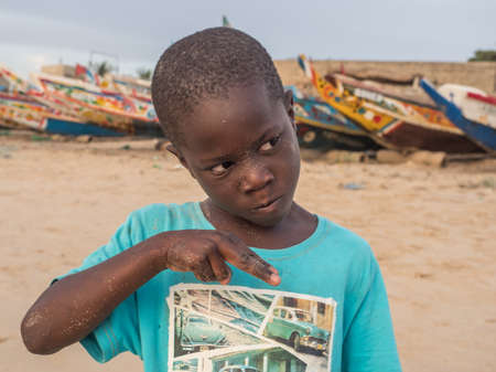 Senegal, Africa - January 24, 2019: Portrait of a black boy with a funny face. Colorful Senegalese boats in the background. Senegal Africa.のeditorial素材