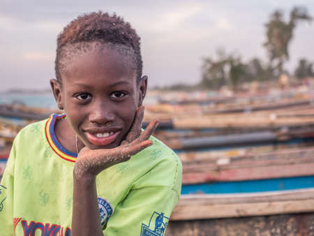 Senegal, Africa - January 24, 2019: Portrait of a black boy with a funny face. Colorful Senegalese boats in the background. Senegal Africa.のeditorial素材