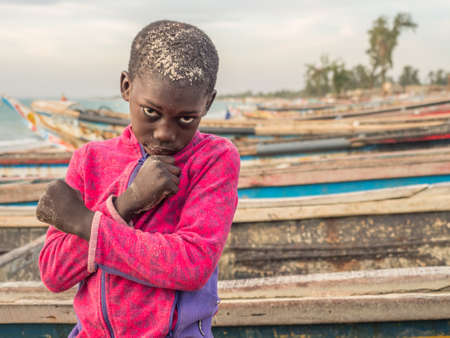 Senegal, Africa - January 24, 2019: Portrait of a black boy with a big eyes. Colorful Senegalese boats in the background. Senegal Africa.のeditorial素材