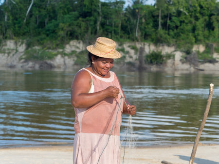 Amazon. Latin America - Sep 2017: Portrait of a happy woman in a hat, a local inhabitant of the Amazon rainforest - a unraveling fishing web on a sandy beach in the Javari Valley. Brazil (Valle del Yavari)のeditorial素材