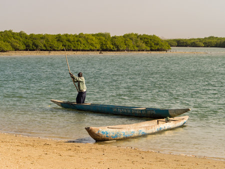 Joal-Fadiouth, Senegal, Africa - Jan 2019: An African man pushes himself off with a pole on a small wooden canoe in the sea between Joel and Fadiouth - next to the island of seashells.のeditorial素材