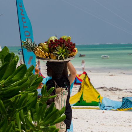 Paje, Zanzibar, Tanzania - January 202: African man with a tray full of exotic fruits on his head. Surfing beach in Paje in the background, Africaのeditorial素材