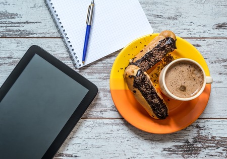 Coffee, cake, eclair with chocolate, tablet, notebook and pen close-up on vintage wooden background. Top view.の写真素材