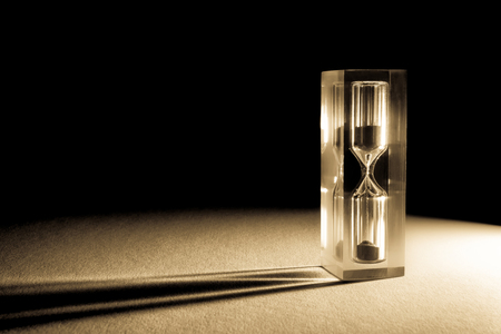 Hourglass sandglass with shadows and glare of the sun on a dark background. Old retro style vintage photo. Sand texture, falling sand.の写真素材