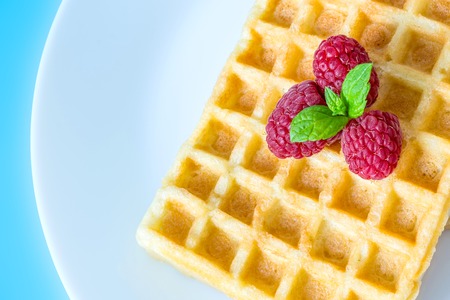 Sweet toast waffles with raspberries and a sprig of mint leaves on a white plate close-up macro on a blue backgroundの写真素材