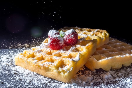 Sweet toast waffles with raspberries and a sprig of mint leaves on top and sifting pouring sugar powder in the sunlight close-up macro on a black backgroundの写真素材