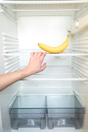 A man's hand reaches out for a single banana in an open, empty refrigerator. Weight loss diet concept.の写真素材