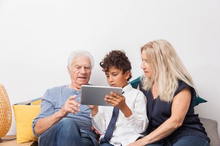 Authentic moment when boy with her grandmother and his grandfather play with a digital tablet on sofa. Smiling family with tablet at home.の写真素材