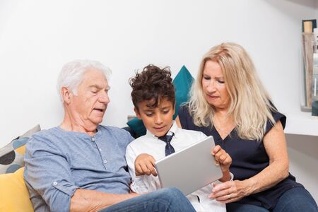 Authentic moment when boy with her grandmother and his grandfather play with a digital tablet on sofa. Smiling family with tablet at home.の写真素材