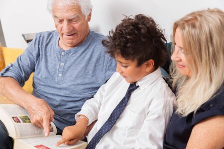 Loving grandpa and grandma with grandson reading together a dictionnary on sofa. Boy reading a book with grandparents.の写真素材