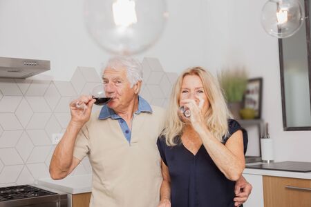 Happy senior couple relax talking and drinking red wine glasses together in the kitchen at home.の写真素材