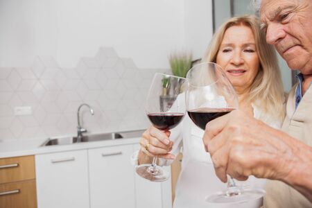 Happy senior couple relax talking and drinking red wine glasses together in the kitchen at home.