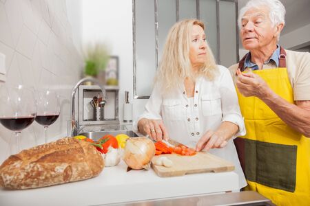 Happy senior couple cooking together with healthy food in a kitchen at home. Blonde woman cutting vegetables.の写真素材