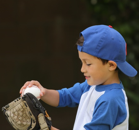 Young cute hispanic boy playing with baseball and glove wearing blue hat on dark backgroundの写真素材