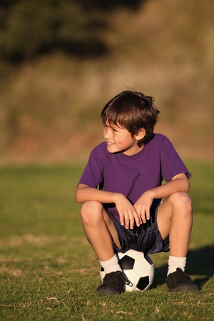 Boy sits on soccer ball looking to side in late afternoon の写真素材