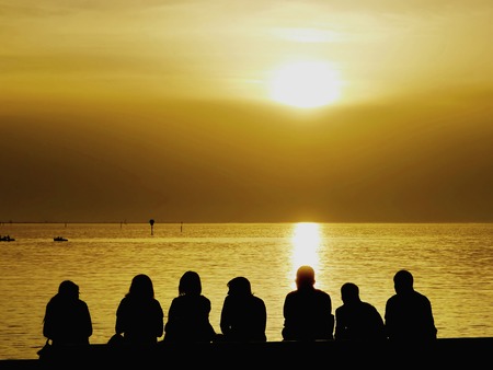 The rear view of the silhouettes of a group of tourists sitting on the seashore, watching a spectacular sunset with light reflected on the surface of the seaの写真素材