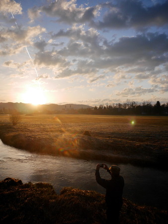 Back view of traveller taking photo of sunset in cloudy sky in autumnal forest while standing near the river.の写真素材