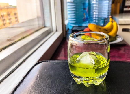 A glass of absinthe with burnt sugar and ice stands on a stand near the window of the apartment.