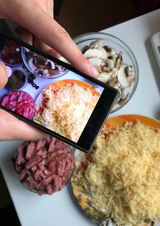 Shooting on a smartphone while cooking pizza at home. Man takes on the smartphone ingredients for pizza: mushrooms, cheese, sauce and sausageの素材