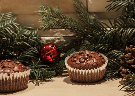 Two chocolate chip cupcakes in paper, a red christmas bell, a pine cone and some green pine branchesの写真素材
