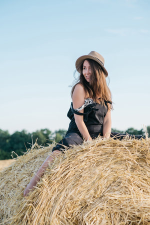 beautiful brunette woman in black overalls with a straw hat on a haystack in a field at sunset