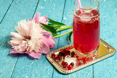 Pink peony and cold homemade drink with cranberry juice and ice in a glass with a cocktail straw on mirror metal plate on a blue wooden table. Selective focusの写真素材
