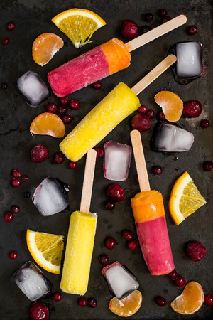 Delicious fruit ice pop with orange, mandarin, cranberries and cherry on old dark iron textured background. Top view, verticalの写真素材