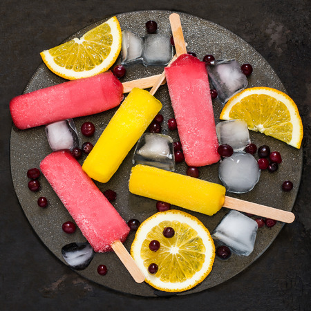 Delicious fruit ice pop with orange and cranberries and ice on a glass tray at the old iron textured background. Top view, square imageの写真素材