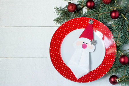 The idea of Christmas decoration for the table setting. Homemade decor for serviette in form of Santa Claus on a plate. Decorated Christmas tree branch on a white wooden tableの写真素材