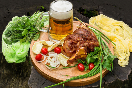 Picnic. Tasty fried on grill pork knuckle, mug with beer, green and pickled onion, parsley, tomatoes, potato chips on large round board. Board with products and colored gauze napkin are on old stumpの写真素材