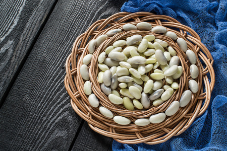 White beans on round wicker tray, blue gauze napkin on dark wooden table. New harvest. Ingredient in...