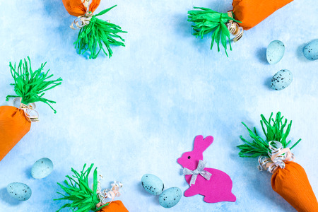 Packs in form of carrots from crepe paper with candy, Easter bunny and decorative eggs on light blue background. Original frame from easter attributes of handworkの写真素材