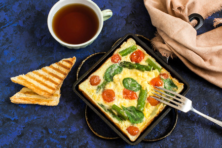 Omelette with asparagus, cherry tomatoes and spinach. Tasty breakfast. Frittata in cast-iron frying pan, toast and cup of tea. Top viewの写真素材