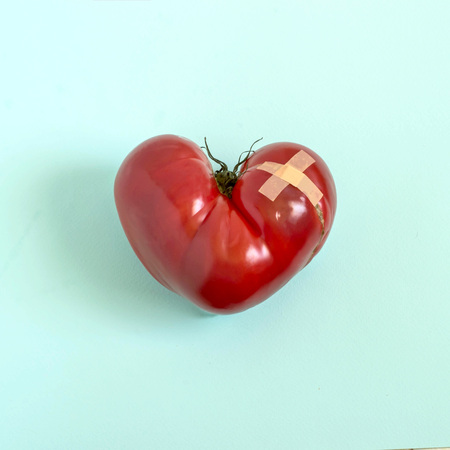 Big tomato in shape of wounded heart with adhesive plaster on blue pastel background. Minimal style. Jokes of nature, imagination and fantasy. Foods useful for heartの写真素材