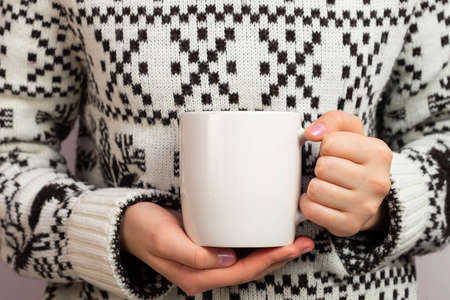 Mockup of white mug in hands of girl wearing sweater. Empty mug mockup for presentation and design promotionの写真素材
