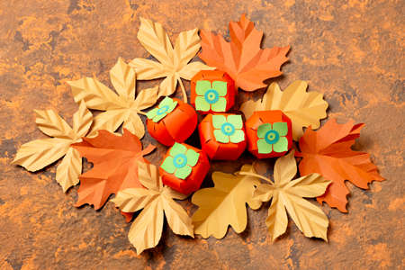 Paper realistic persimmon and autumn leaves on brown textured background. Paper art and craft. Trendy hobby. Autumn food art conceptの写真素材