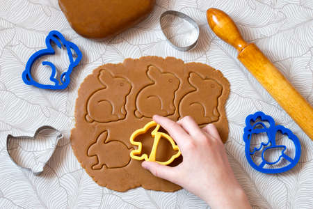 Cutting out Easter gingerbread in shape Easter bunnies from dark honey dough with spices. Dough, rolling pin and different Easter cookie cutters on tableの写真素材