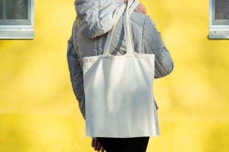 Empty reusable canvas tote bag mockup. Natural canvas eco-friendly shopper bag. Mockup for presentation of design or brand. Teen girl stands at yellow wall of house and holds tote bag on her shoulderの写真素材