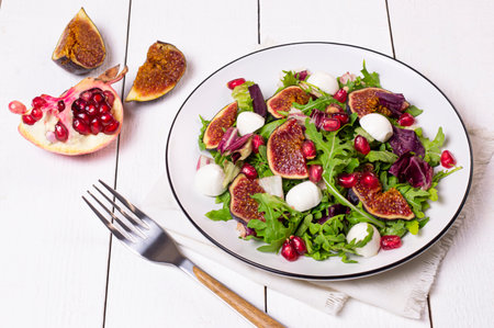 Delicious salad with figs, mozzarella, arugula, radicchio and pomegranate seeds on white wooden background. Concept of diet and healthy foodの写真素材