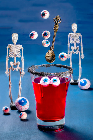 Glass of Halloween cocktail with eyeballs surrounded by skeletons. Eyeballs and spoon flying into glass. Scary drinks with decorations for Halloween partyの写真素材