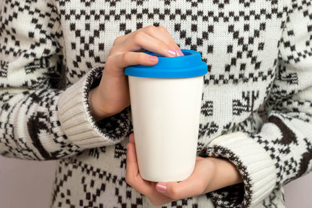 Mockup of blank white ceramic travel cup with blue lid in hands of girl dressed in sweater. White coffee reusable cup for presentation and promotion designの写真素材