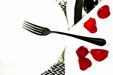 black silverware on white plate and folded napkin with loose red rose petalsの写真素材