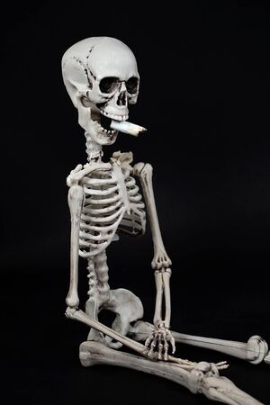 skeleton in sitting position with cigarette butt in mouth isolated on blackの写真素材